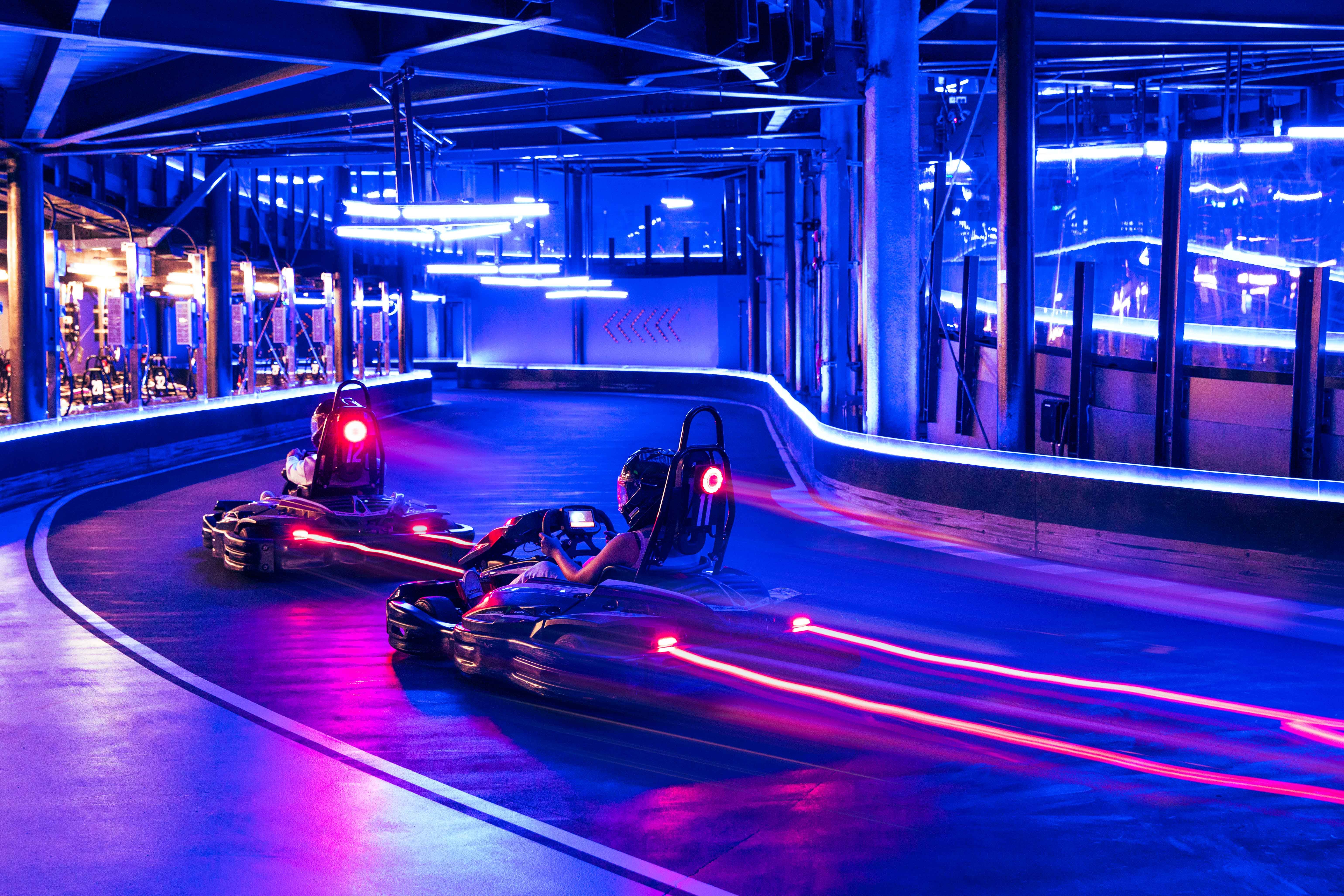 Two go-karts racing on an indoor neon-lit track with motion trails and glowing blue and pink lights